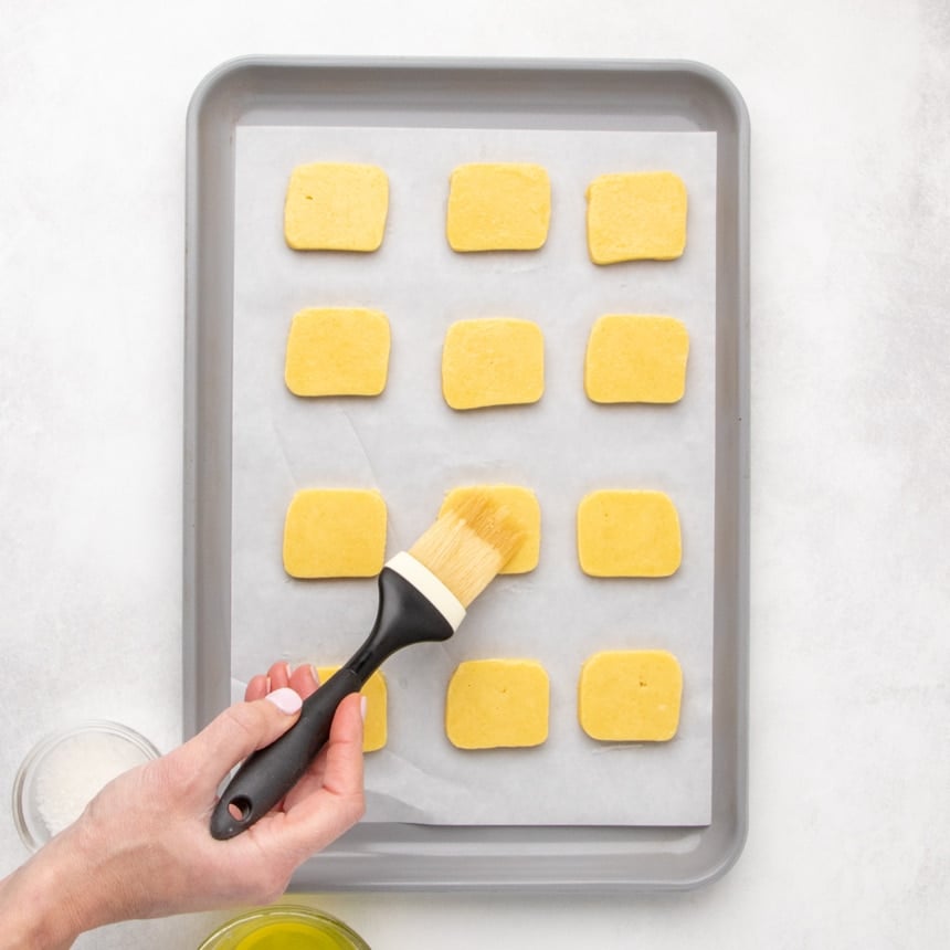 A hand brushing the top of one small flat rectangular dough piece on a parchment-lined baking sheet; several evenly spaced dough pieces are arranged in rows.