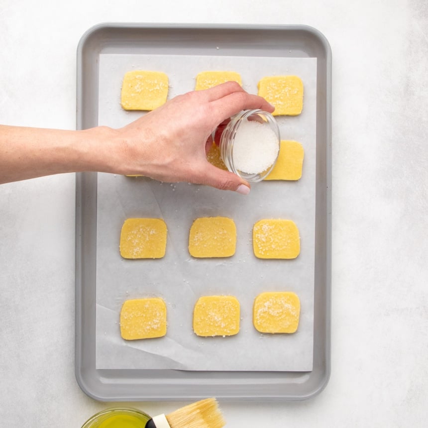 A hand sprinkling coarse white sugar from a small glass bowl over dough pieces on a parchment-lined baking sheet.