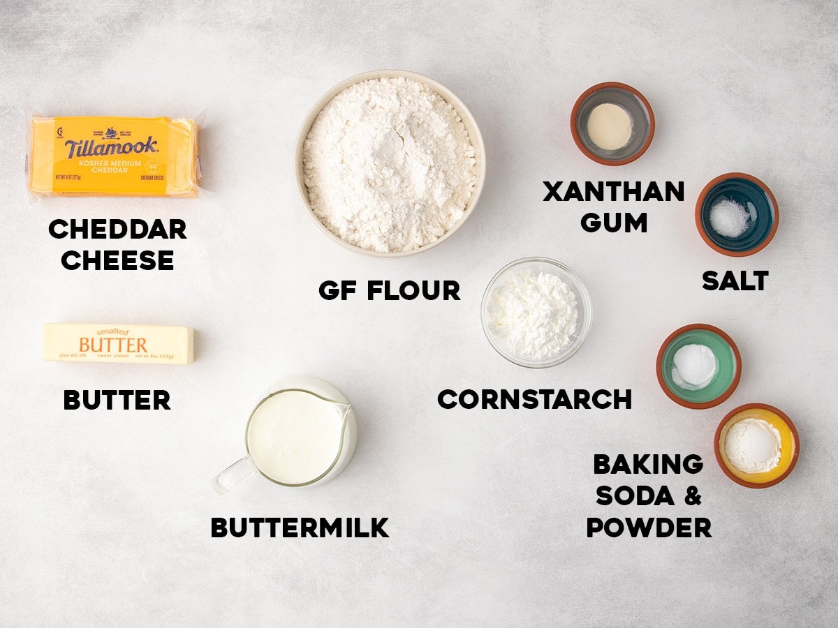 Measured ingredients for gluten free cheese scones are arranged on a light surface, including cheddar cheese, gluten free flour, xanthan gum, salt, butter, cornstarch, buttermilk, and baking soda and powder, each labeled with text.