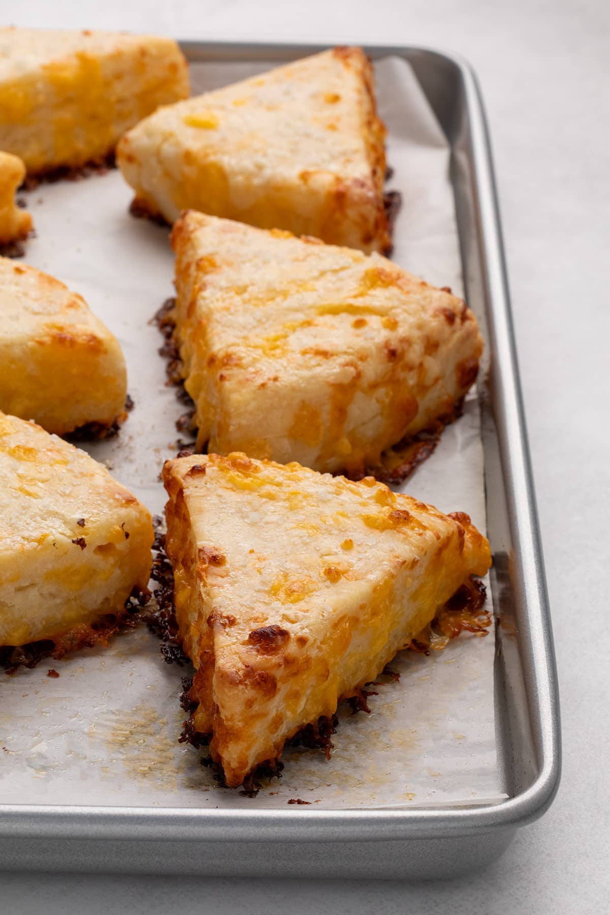 Baked triangular cheese scones with melted cheese are shown from a side angle on a parchment-lined baking sheet.