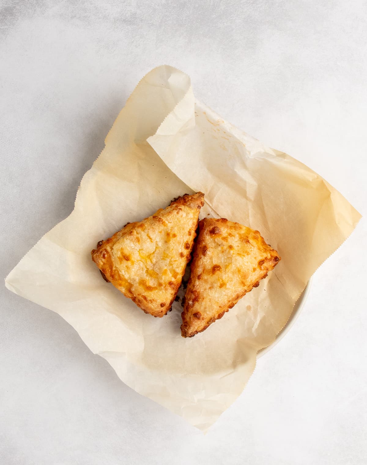 An overhead view shows baked triangular cheese scones spaced on a small round plate barely visible beneath lightly browned parchment paper.