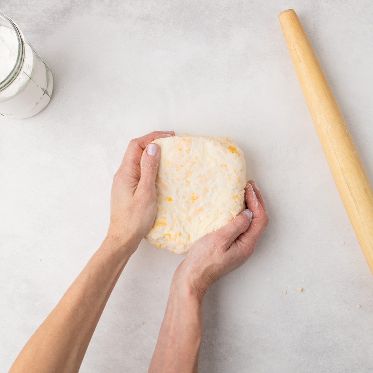 Two hands gently shape the same raw dough into a compact block on the floured surface, with a rolling pin off to the side.