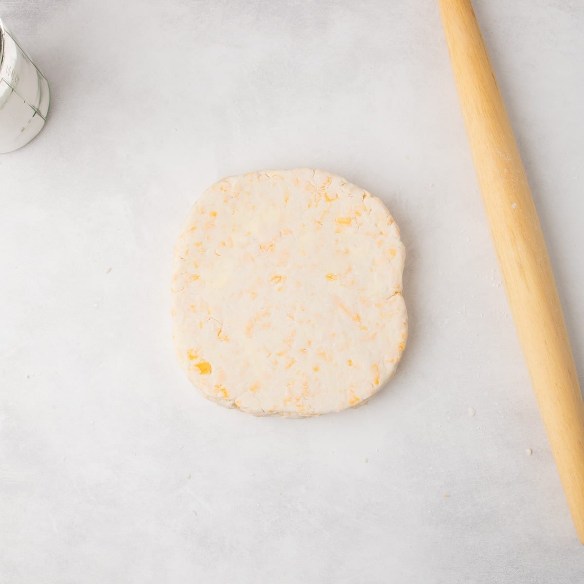 A round disk of raw cheese scone dough with visible bits of cheese sits on a floured surface, with a rolling pin nearby.