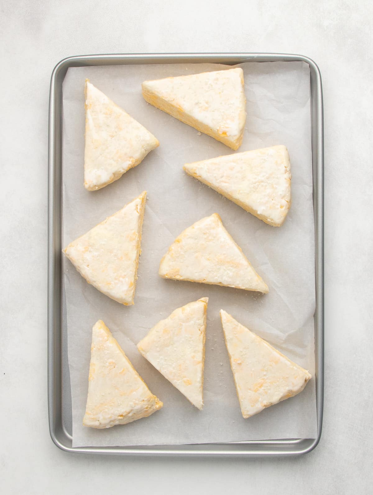 Unbaked triangular pieces of raw dough are spaced apart on a parchment-lined metal baking sheet.