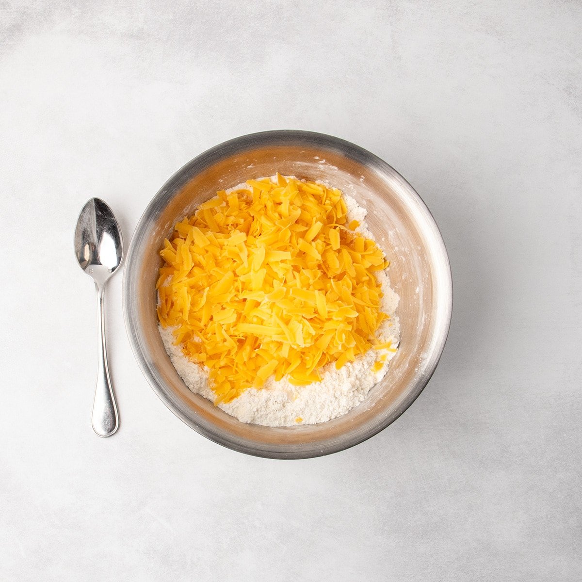 Shredded orange cheese is piled on top of flour in a metal mixing bowl; a metal spoon rests beside the bowl.