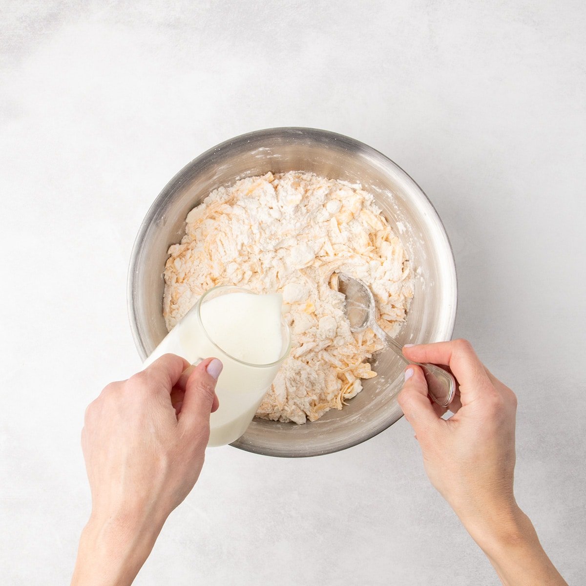 Milk is being poured from a measuring cup into a metal bowl of flour, butter shards, and shredded cheese while a spoon is held inside the bowl.