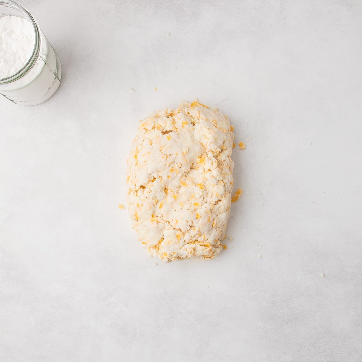 A rough mound of raw cheese scone dough with visible shreds of orange cheese sits on a lightly floured surface; a glass jar of flour is nearby.