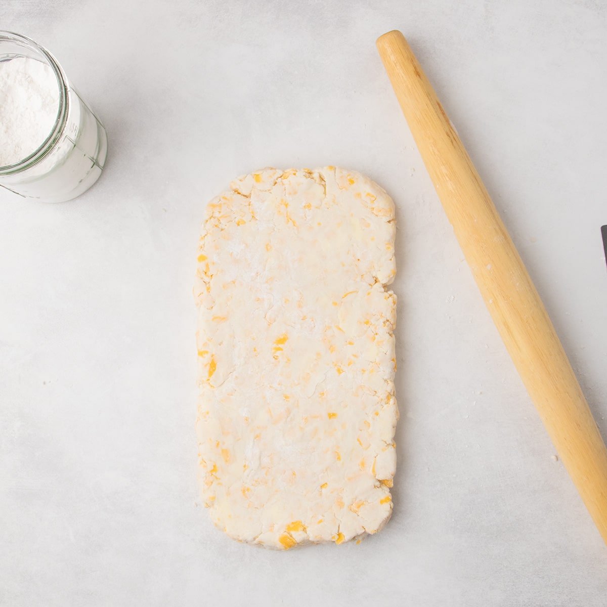 The raw cheese scone dough is flattened into a rectangular shape on a floured surface, with a rolling pin placed beside it.