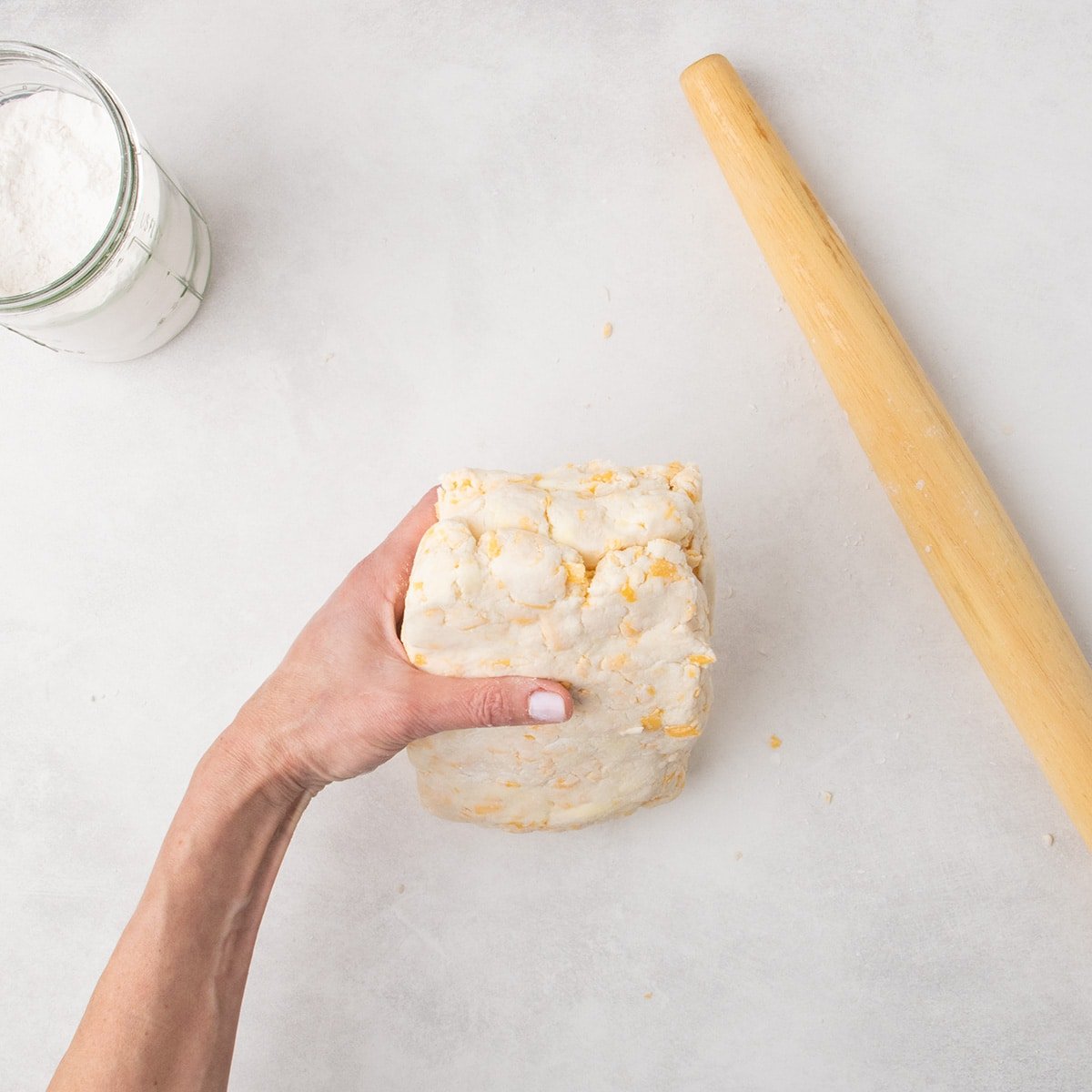 A hand stacks three pieces of raw dough on top of another, forming a thicker block on the work surface.