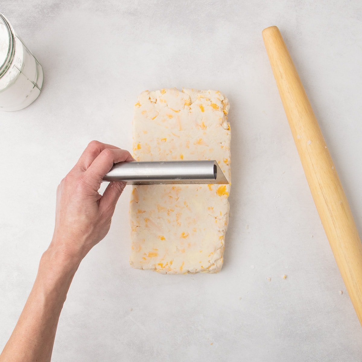 The stacked dough is pressed into a rectangular shape again, with a metal bench scraper held above it.