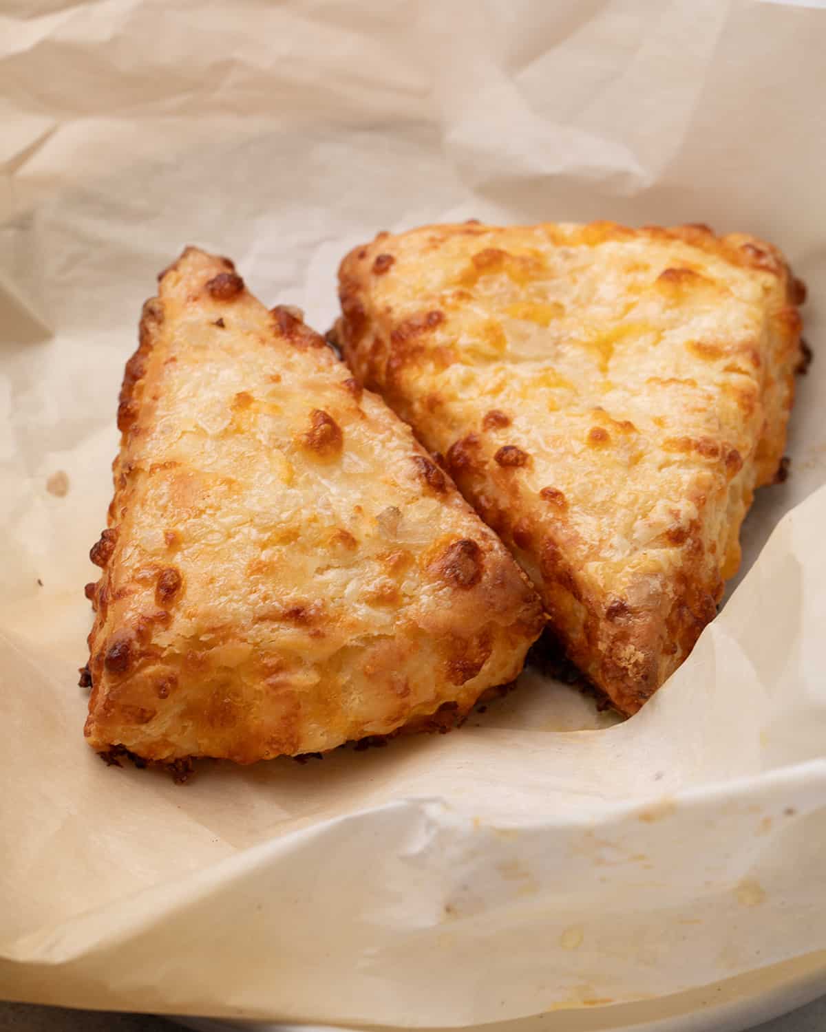 Two baked triangular gluten free cheese scones rest on a sheet of parchment paper on a plate.