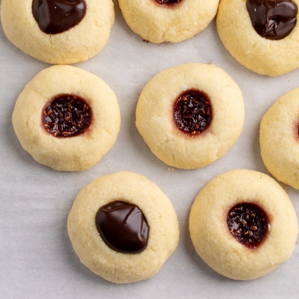 Close-up view of baked gluten free thumbprint cookies on parchment paper, some filled with chocolate and others with a dark red jam.
