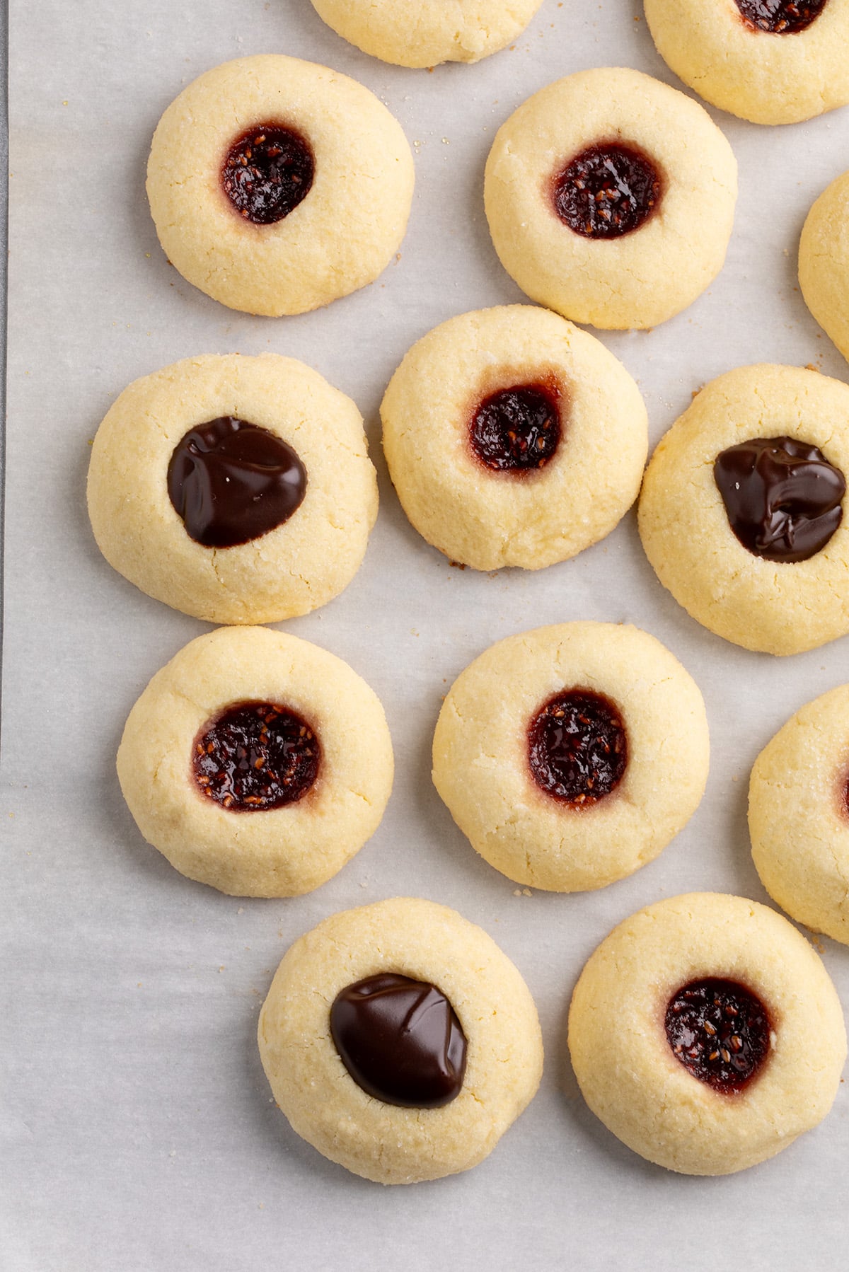 A larger group of thumbprint cookies arranged on parchment paper, with some filled with chocolate and others with a dark red jam.