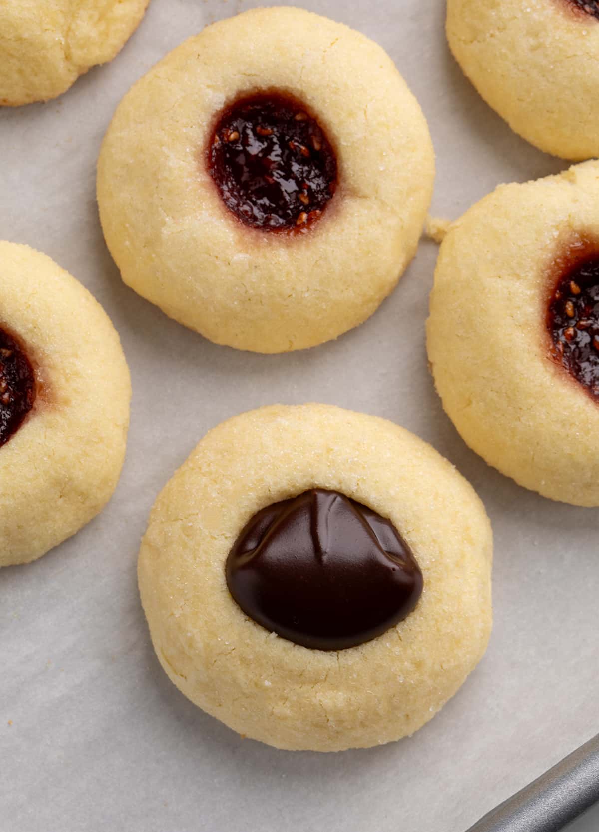 Close-up of several thumbprint cookies on parchment paper, showing both chocolate-filled centers and dark red jam-filled centers.