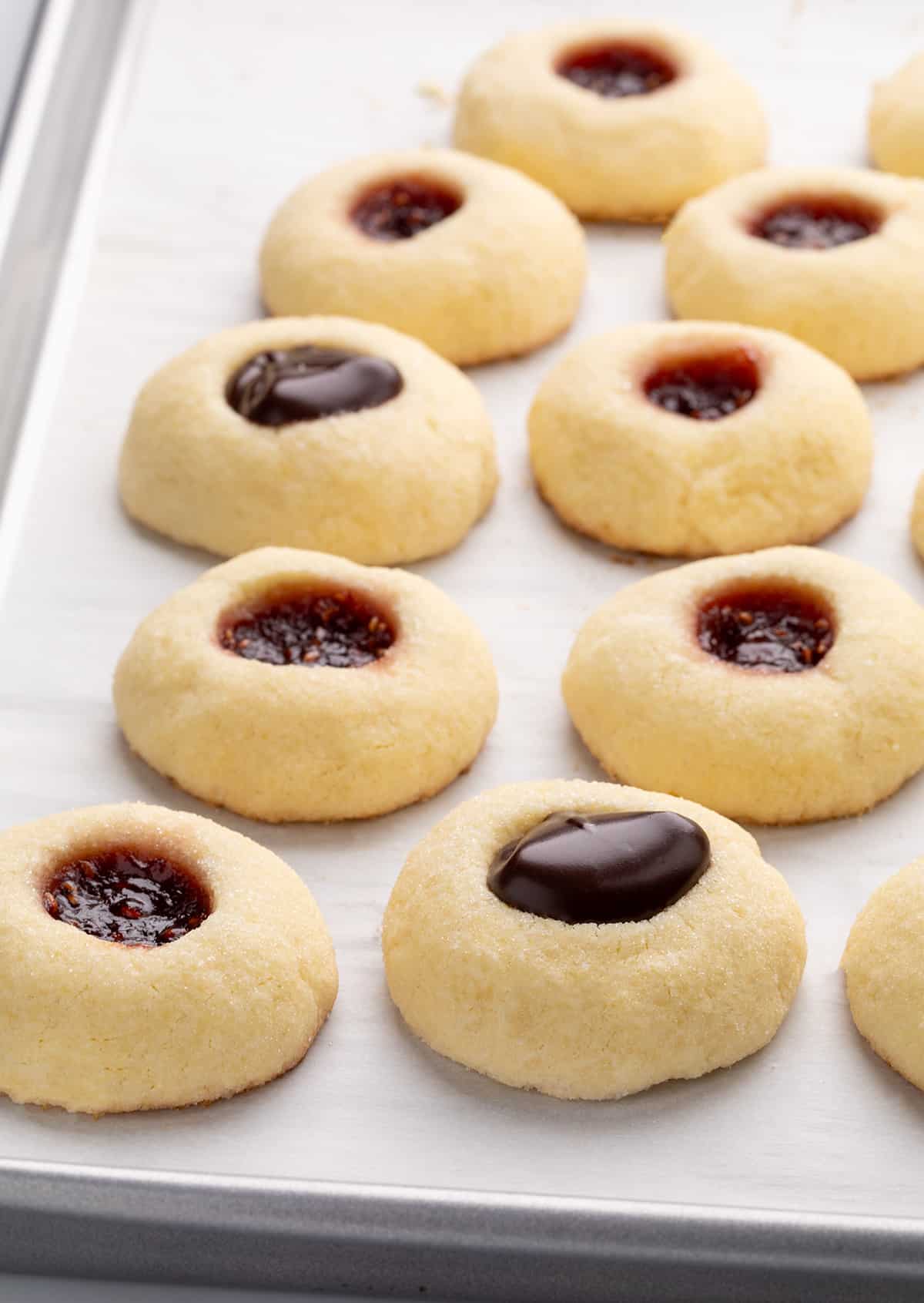 Side-angle view of thumbprint cookies on a parchment-lined baking sheet, with some filled with chocolate and others filled with dark red jam.