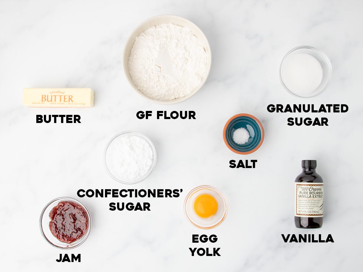 Overhead view of labeled baking ingredients arranged on a light marble surface, including butter, gluten free flour, granulated sugar, salt, confectioners’ sugar, jam, an egg yolk, and a bottle of vanilla extract.