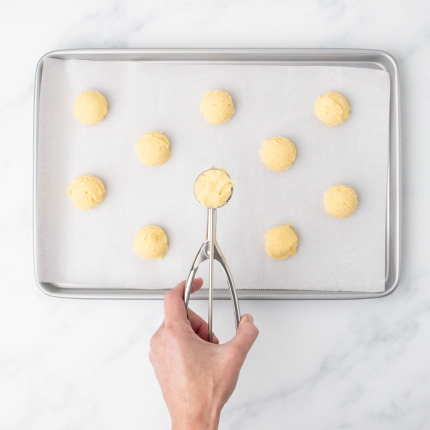 A hand holding a small metal cookie scoop above a parchment-lined baking sheet with evenly spaced scoops of light yellow dough.