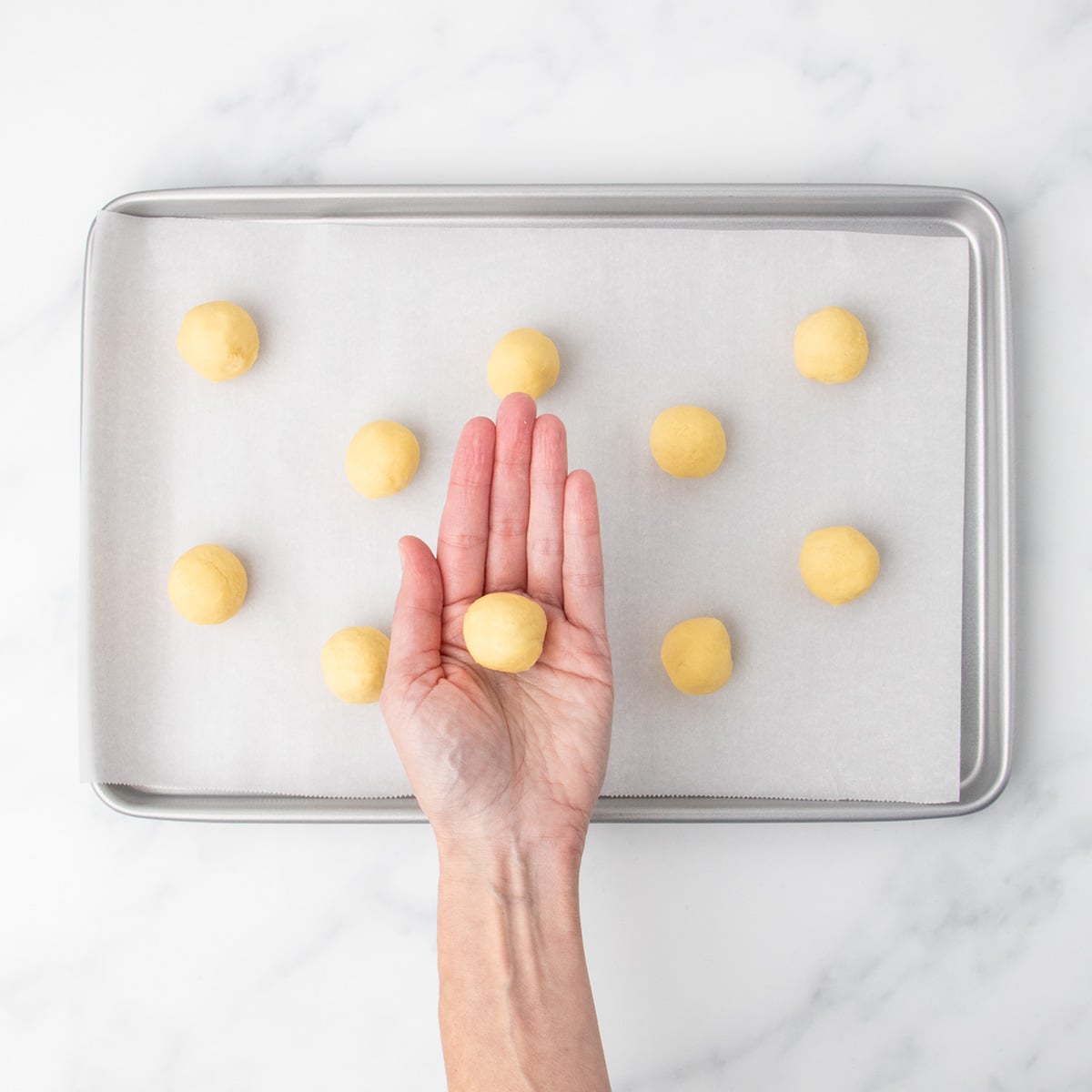 A hand holding a round ball of light yellow dough above a parchment-lined baking sheet with more dough balls arranged on it.