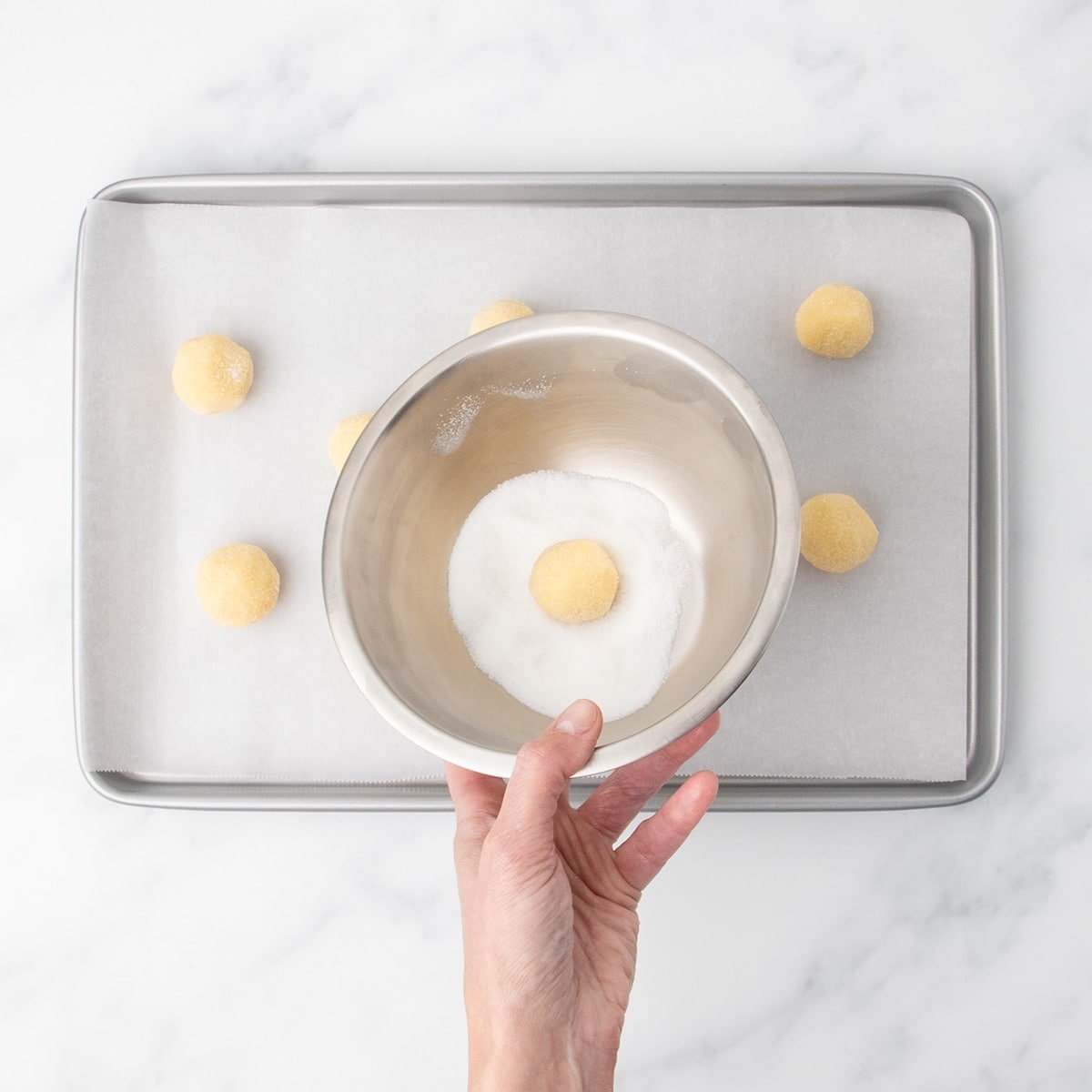 A hand holding a small metal bowl filled with granulated sugar, tilting a dough ball in the bowl to cover it in granulated sugar.