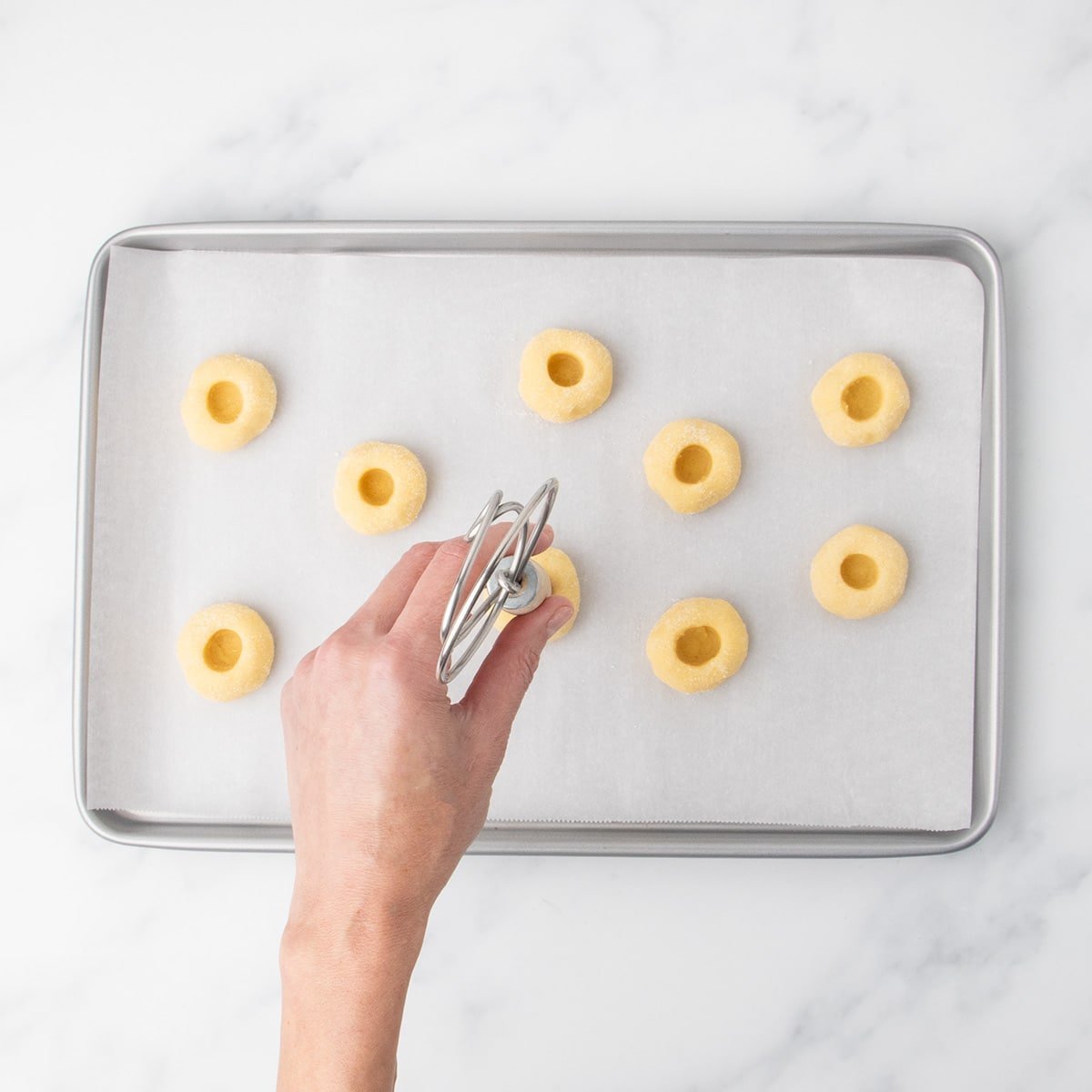 A hand pressing an indented center into dough balls on a parchment-lined baking sheet using a metal whisk end.