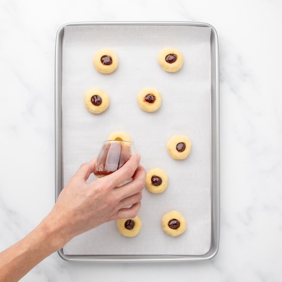 A hand holding a small glass container pouring slightly warmed jam filling into the indents of the dough balls.
