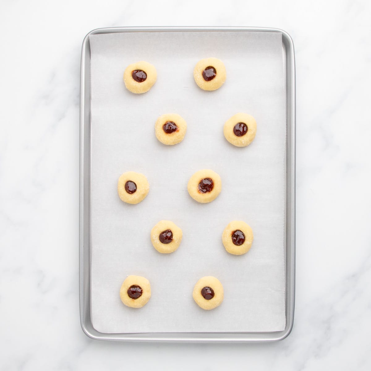 A baking sheet lined with parchment paper holding 10 raw dough balls with jam filling in the centers before baking.