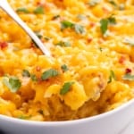 A spoon lifting a portion of baked gluten free macaroni and cheese from a white oval baking dish, with melted cheese and green leaves on top.