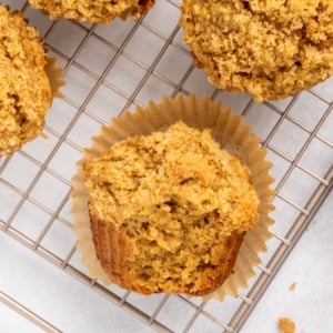 Closeup of a single baked muffin in a paper liner on a wire cooling rack, with a crumb-topped surface and a broken section revealing the interior, with other whole muffins in above and to the side.
