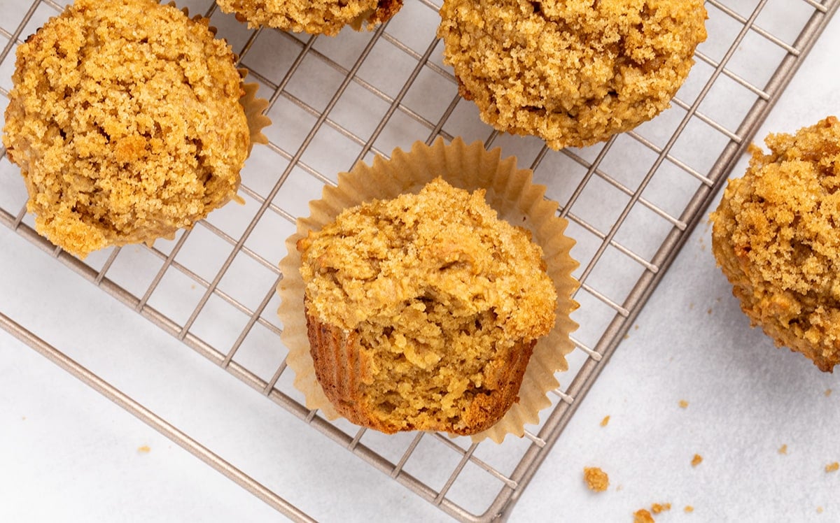 Several baked muffins in paper liners arranged on a wire cooling rack, viewed from above, with one on its side with a bite taken, revealing a tender interior.