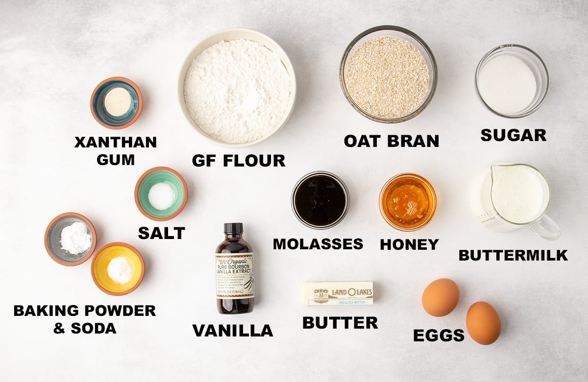 Measured baking ingredients arranged on a light marble surface, including bowls and containers labeled xanthan gum, gluten free flour, oat bran, sugar, salt, baking powder and soda, molasses, honey, vanilla, butter, buttermilk, and eggs.