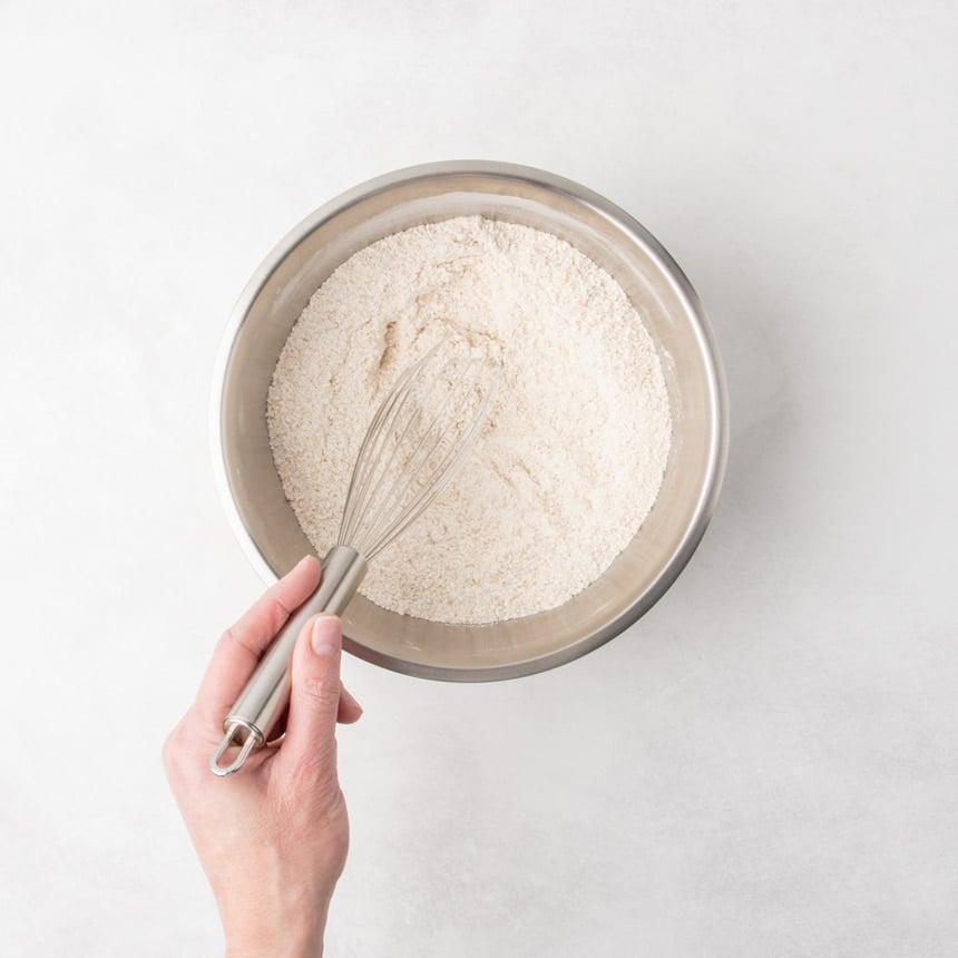 A hand holding a whisk, mixing dry ingredients in a stainless steel bowl.