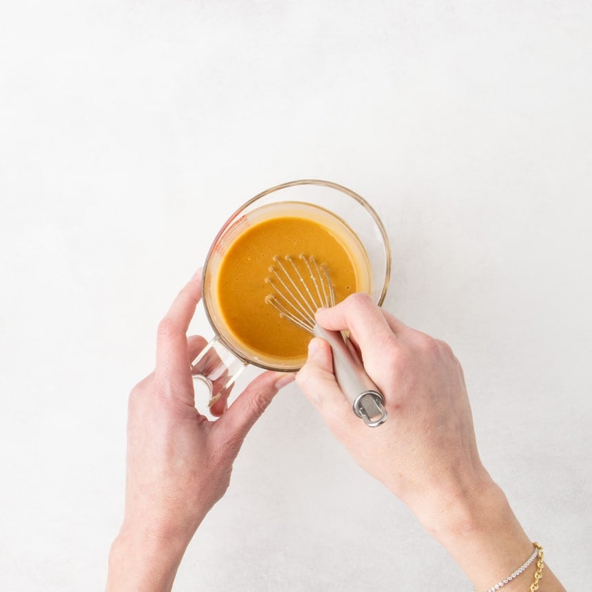 Two hands whisking a brownish orange-colored liquid mixture in a clear measuring cup.
