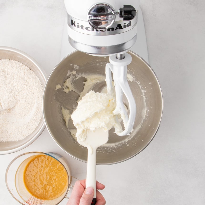 Stand mixer bowl with a paddle attachment holding a thick white mixture of whipped butter and sugar, with dry ingredients and an orange liquid nearby.