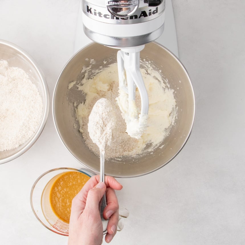 A handheld spoon adding dry ingredients into a stand mixer bowl containing a white mixture.