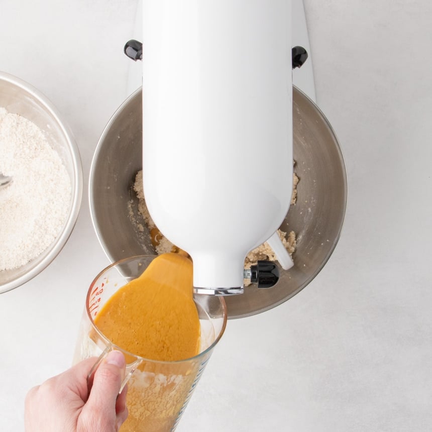 Brownish orange liquid being poured from a handheld measuring cup into a stand mixer bowl with partially mixed batter.