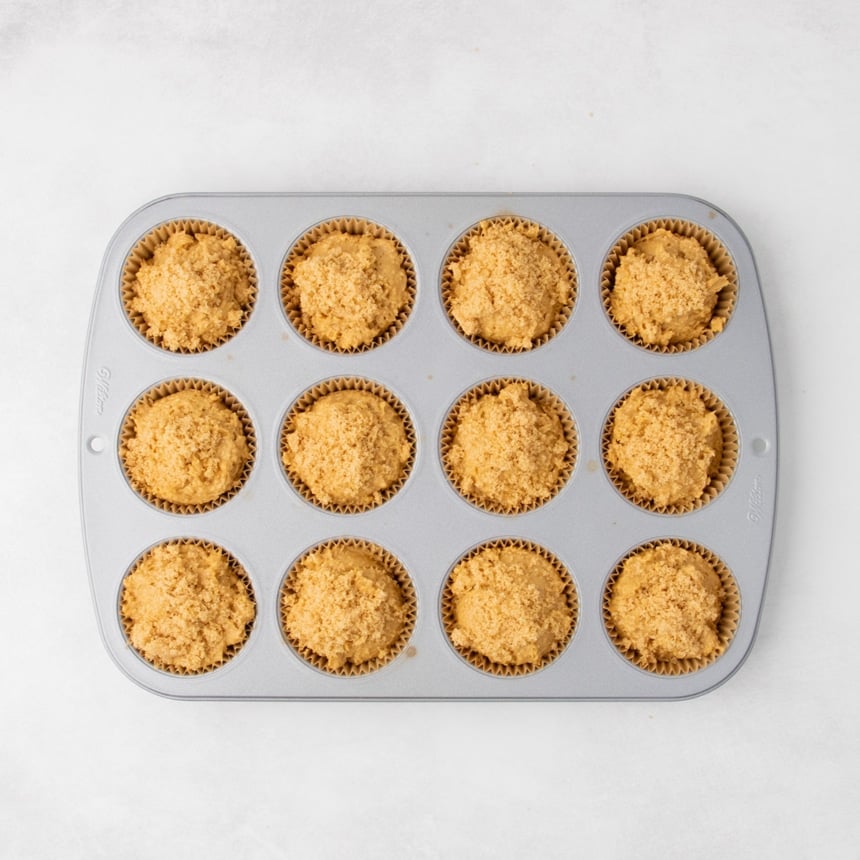 Unbaked muffin batter divided into brown paper liners in a 12-cup muffin pan, topped with a generous sprinkling of light brown sugar.