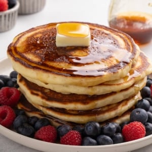 A closeup image of a stack of four browned pancakes topped with a square of butter and syrup, surrounded by blueberries and raspberries on a white plate.