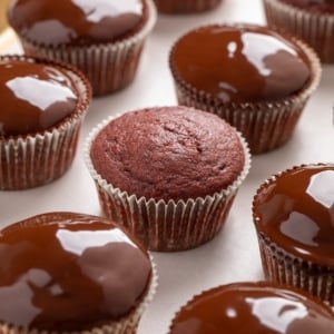 Gluten free red velvet cupcakes in paper liners, with several topped with a glossy chocolate coating and one plain cupcake in the center.