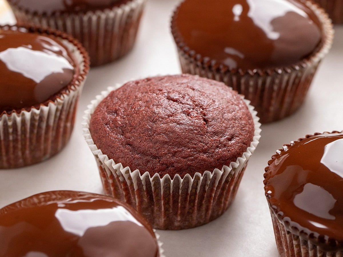 Closeup on a plain gluten free red velvet cupcake in a paper liner surrounded by cupcakes topped with glossy chocolate coating.