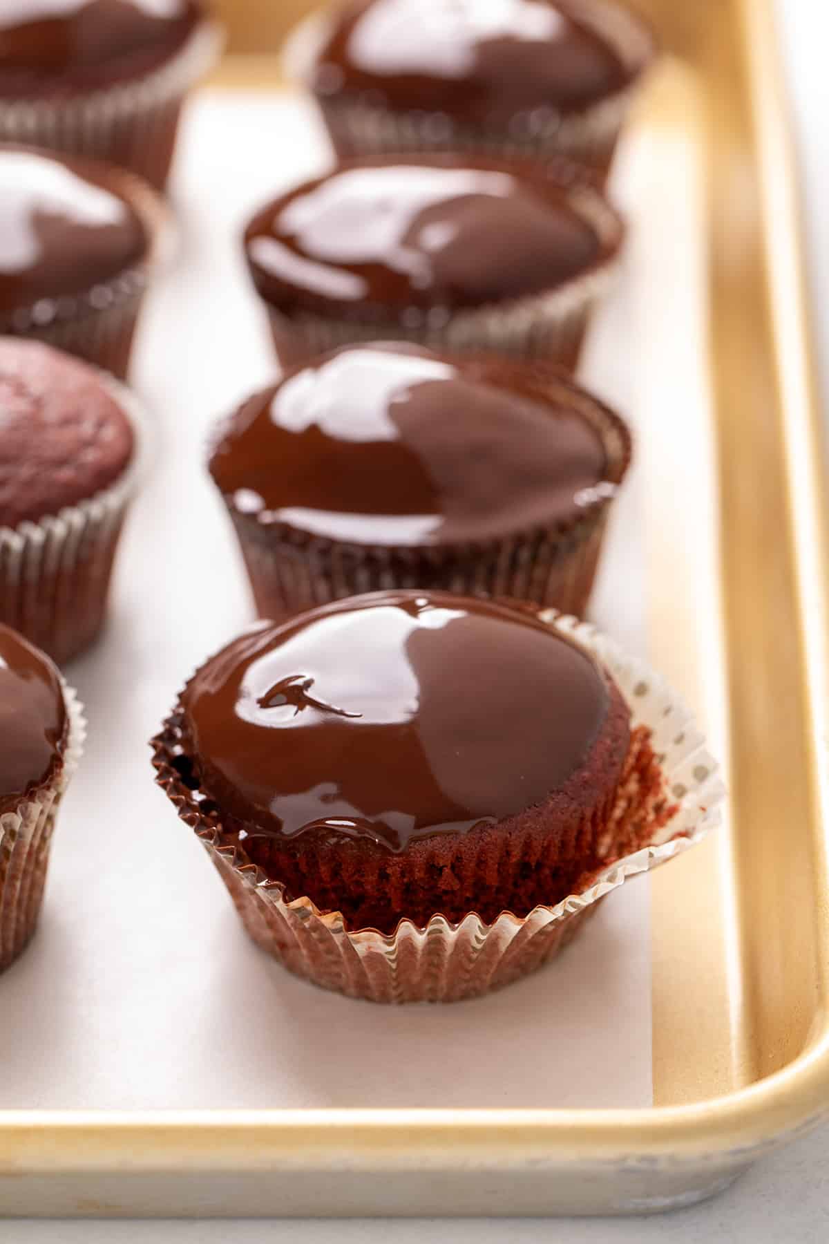 Chocolate-topped red cupcakes arranged on a parchment-lined baking sheet with a gold-colored rim, with the wrapper peeled back from the cupcake in the front revealing the red velvet cake inside.
