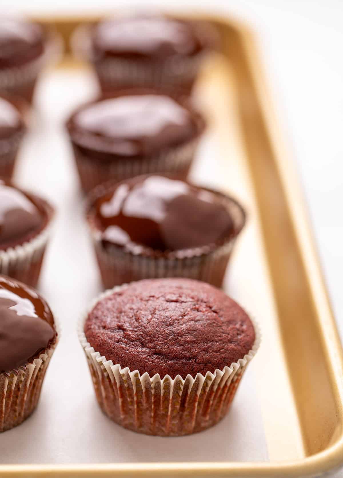 A plain red cupcake in a paper liner on a baking sheet with chocolate-topped cupcakes in the background.