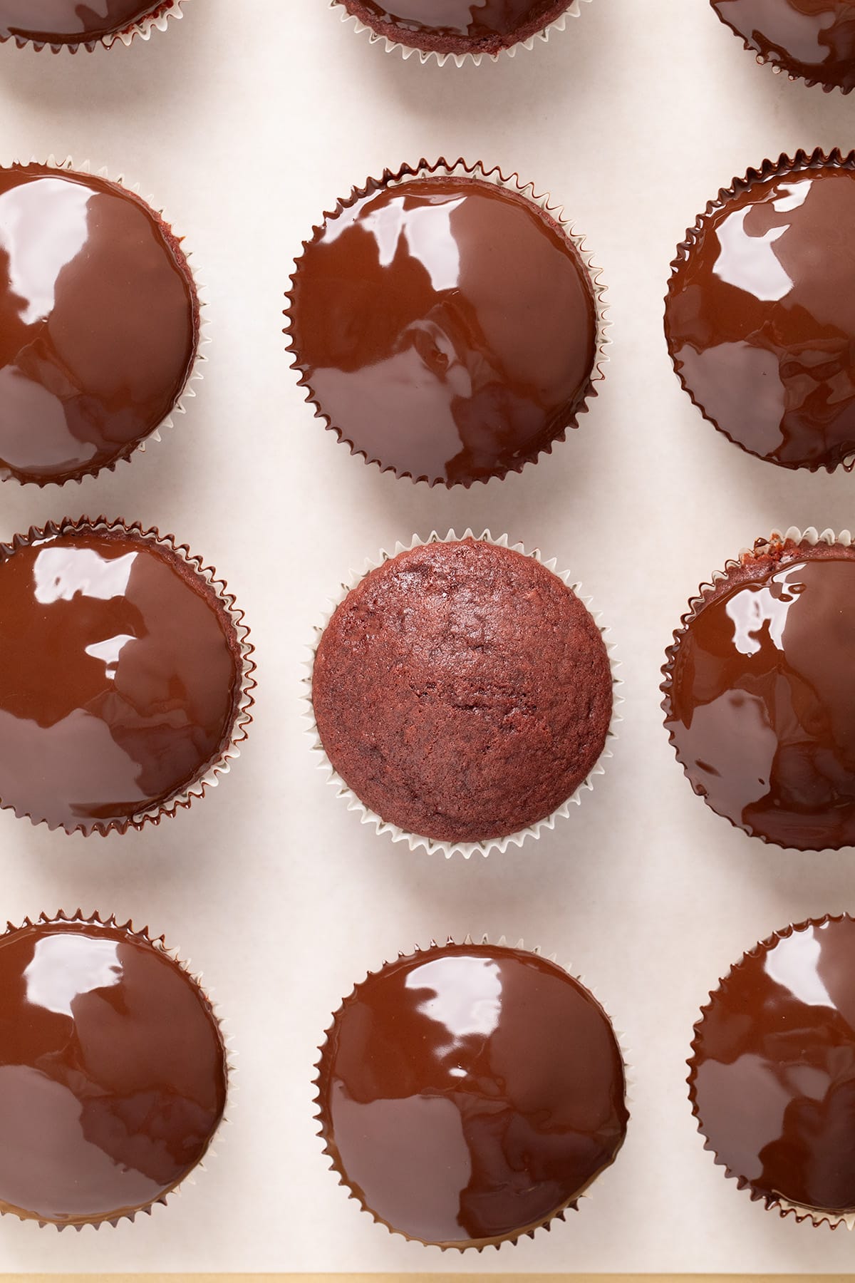 An overhead view of red velvet cupcakes in paper liners, most topped with glossy chocolate coating and one plain cupcake in the center.
