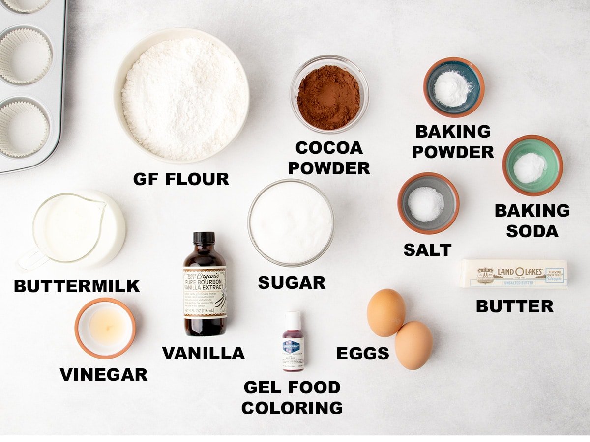 Measured baking ingredients for gluten free red velvet cupcakes arranged on a white surface, each labeled with text, including bowls, eggs, butter, bottles, and a muffin pan.