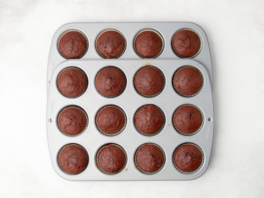 16 baked red cupcakes in paper liners arranged in a stacked metal muffin pans.