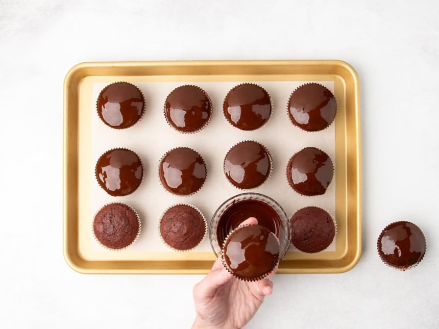 A hand holding a chocolate-topped cupcake above a baking sheet filled with red cupcakes, with a small bowl of chocolate coating nearby.