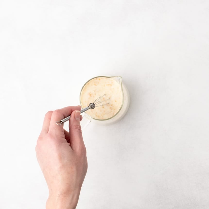 A hand holding a very small whisk stirring a pale liquid mixture in a glass measuring cup on a white surface.