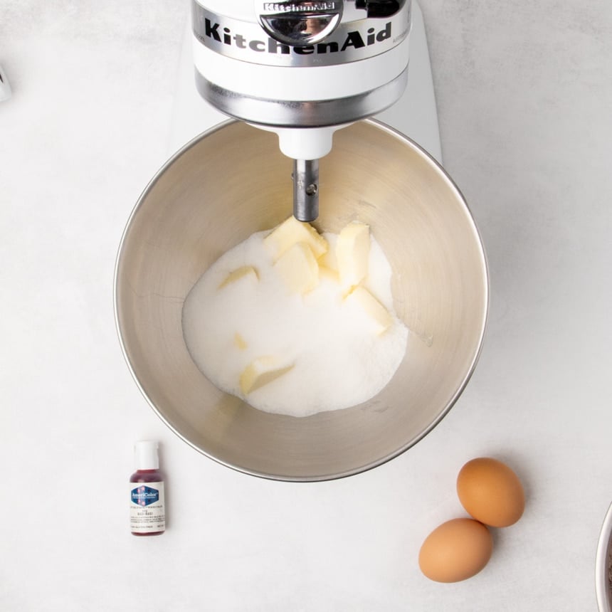 Granulated sugar and chunks of butter in a metal stand mixer bowl beneath a KitchenAid mixer, with two eggs and a small bottle nearby.