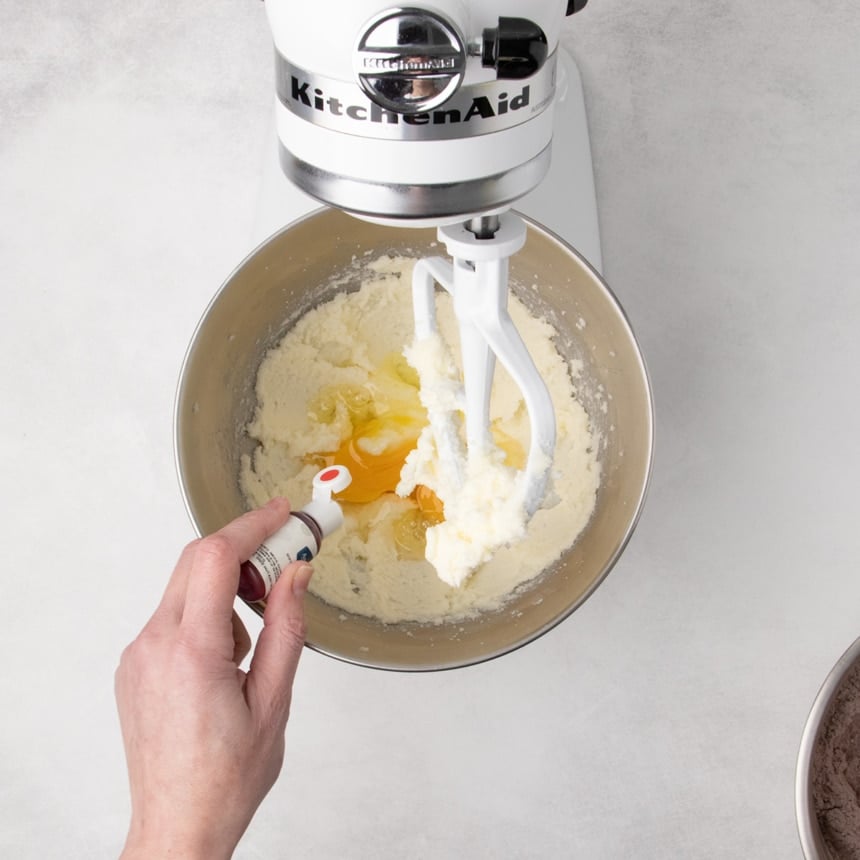 Eggs added to the creamed mixture in a stand mixer bowl, with a hand holding a small bottle above the bowl, about to squeeze in red gel food coloring.