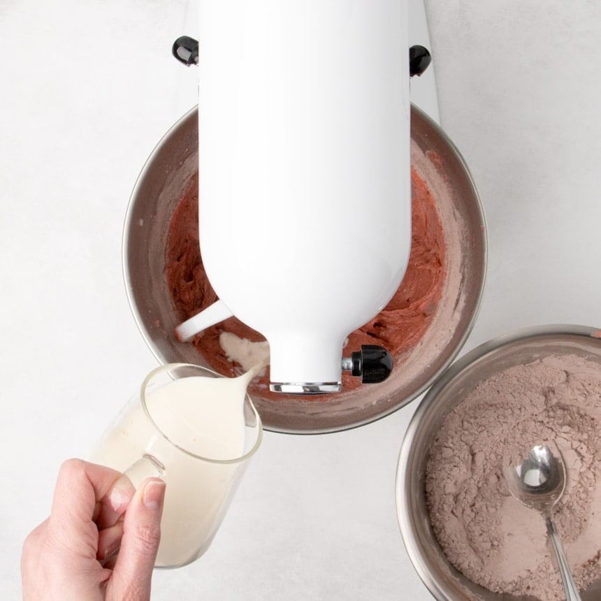 White liquid mixture being poured from a glass measuring cup into a stand mixer bowl containing red batter.