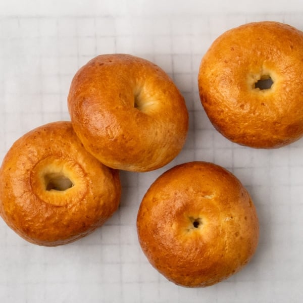 Four golden brown gluten free bagels arranged on a sheet of white parchment paper with a brown-handled bread knife nearby.